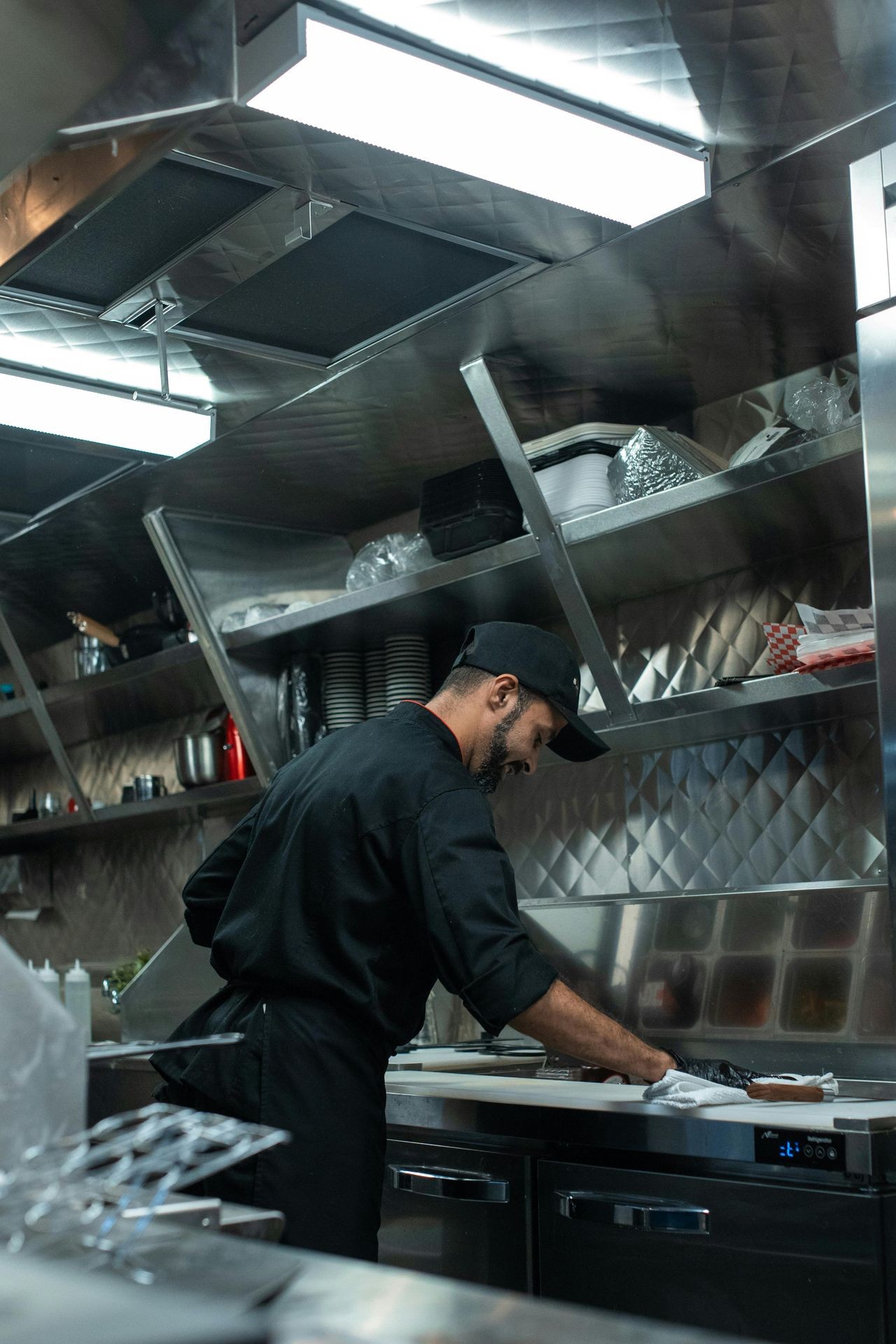 Chef in black uniform working in a commercial kitchen with stainless steel shelves and appliances.