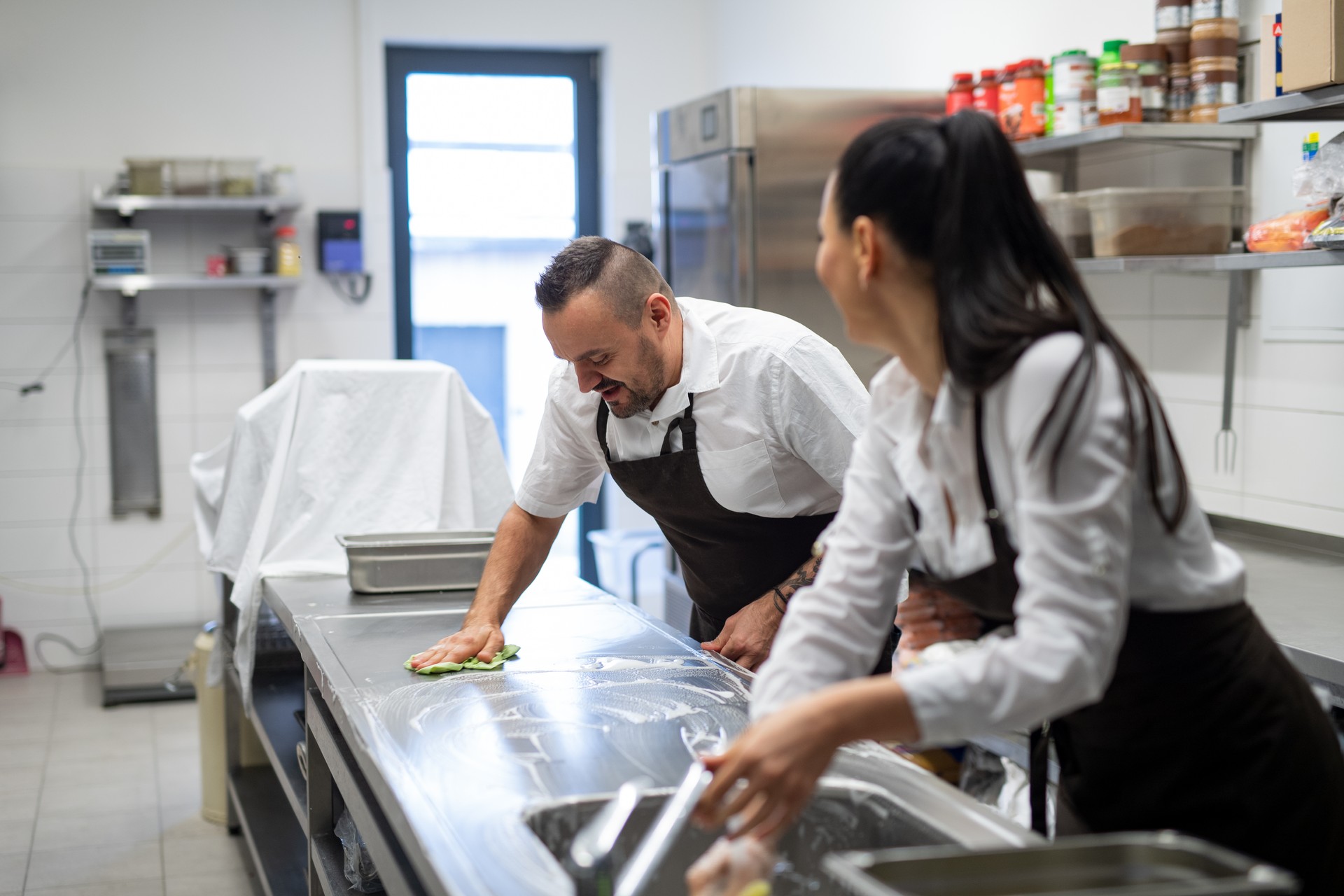 Chef and cook cleaning the workspace after doing dishes indoors in restaurant kitchen.