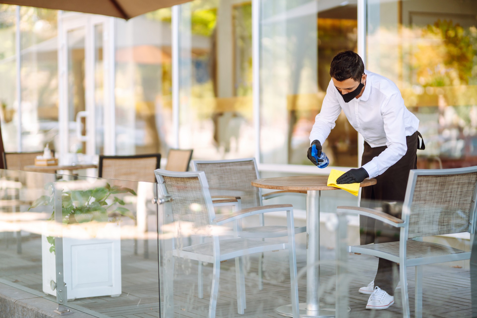 Waiter wearing protective face mask and gloves while disinfecting tables at outdoor cafe.